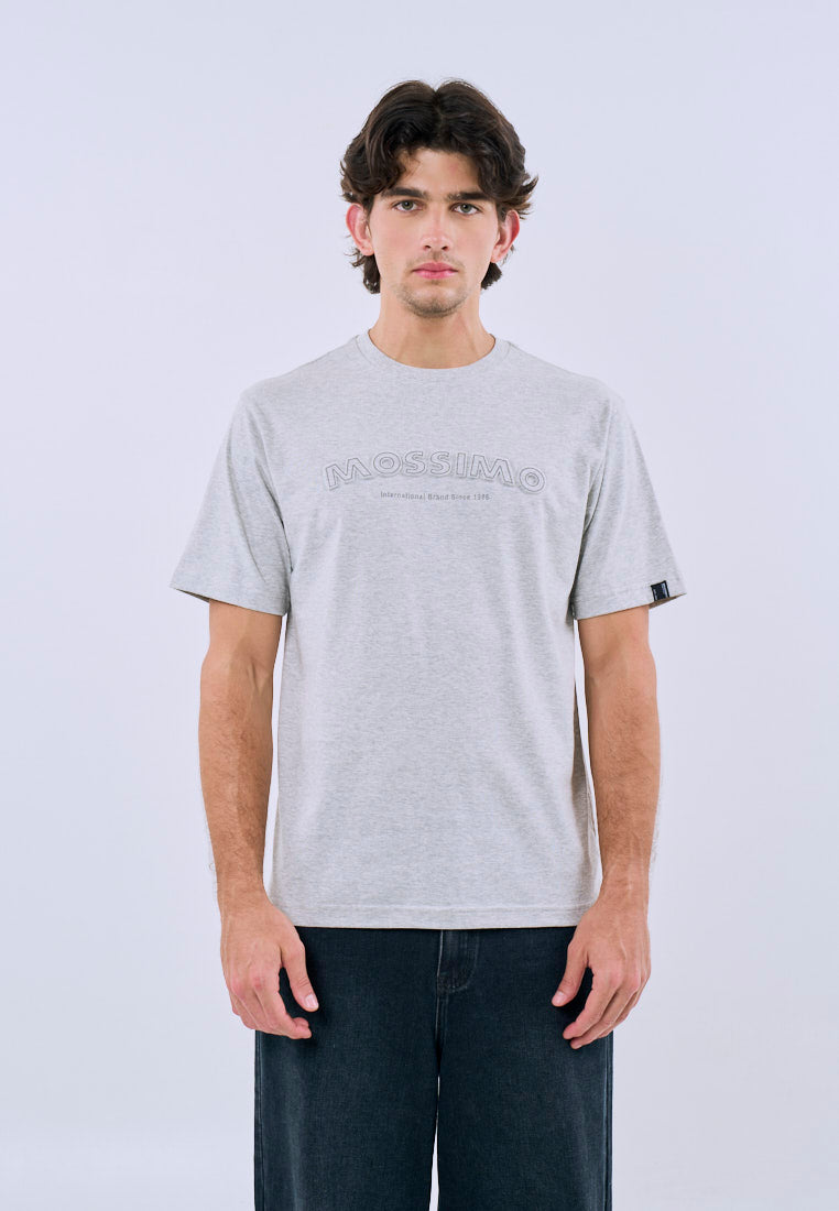 A man wearing a light gray heather crewneck t-shirt with a tonal 'Mossimo' logo across the chest, paired with dark gray jeans, standing against a light gray studio background.